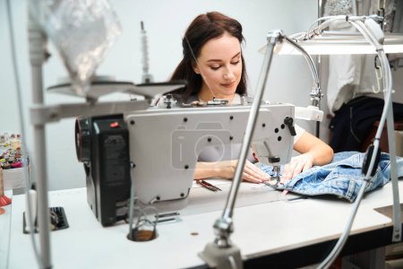 Foto de Etiquetas de costura de costurera concentradas y ropa de procesamiento en una gran máquina de coser industrial, servicio de alteración y confección - Imagen libre de derechos