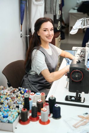 Téléchargez les photos : Femme hautement qualifiée employée de bureau à la blanchisserie assise sur le lieu de travail avec machine à coudre et service de sourire, de modification et de réparation - en image libre de droit