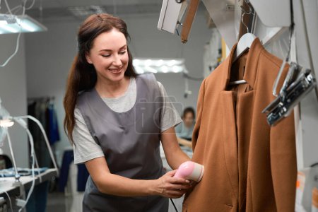 Trabajador de oficina de limpieza en seco mujer calificada usando removedor de pelusa para limpiar el abrigo del cliente, cuidado profesional