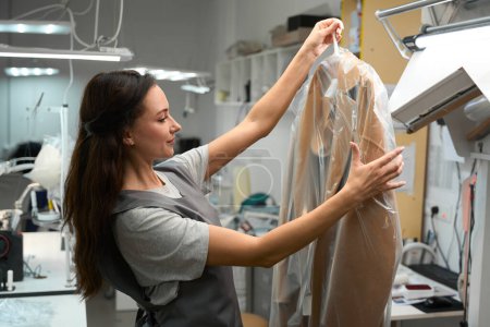 Foto de Mujer trabajadora de oficina de lavandería sosteniendo percha con abrigo seco y limpio en bolsa de celofán, preparándose para llevar al cliente - Imagen libre de derechos