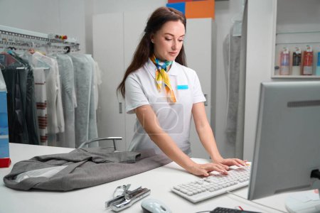 Téléchargez les photos : Opérateur de bureau de blanchisserie femme concentrée tapant sur le clavier, saisissant des données sur les vêtements des clients à programmer - en image libre de droit