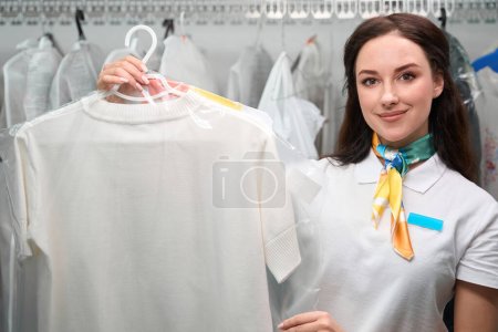 Téléchargez les photos : Femme souriante positive nettoyage à sec employés de bureau tenant cintres avec des t-shirts blancs propres, service de qualité professionnelle - en image libre de droit