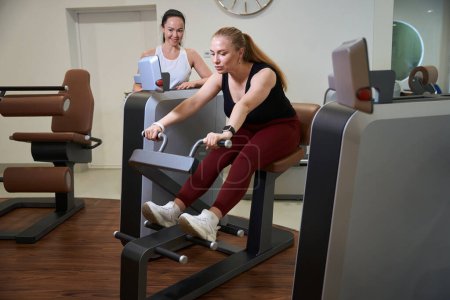 Foto de Hermosa mujer se dedica a una moderna máquina de fitness, junto a un entrenador consultor - Imagen libre de derechos