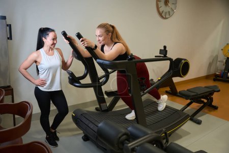 Photo for Female in sportswear is engaged in a simulator, next to a woman is a fitness trainer - Royalty Free Image