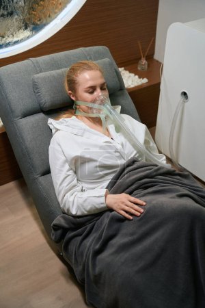 Téléchargez les photos : Femme dans un masque spécial est situé dans une chaise confortable, elle aime la procédure de thérapie hypoxique - en image libre de droit