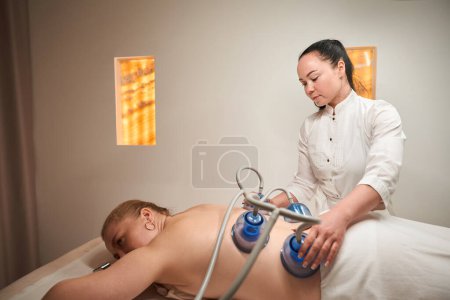 Foto de Rubia se acuesta en la mesa de masaje con su respaldo, la morena realiza el procedimiento de terapia de vacío - Imagen libre de derechos