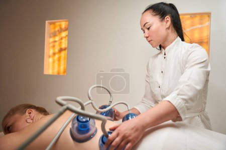 Photo for Young brunette performs a vacuum therapy procedure, the client lies on the massage table with her back up - Royalty Free Image