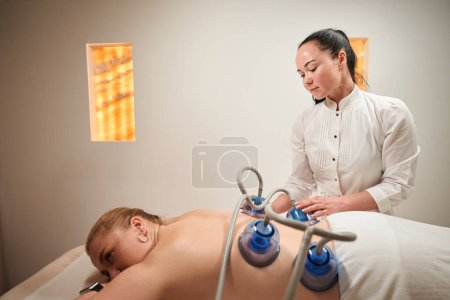 Photo for Blonde lies on a massage table with her back up, a young masseuse performs a vacuum therapy procedure - Royalty Free Image