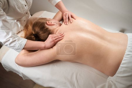 Foto de La masajista femenina hace el masaje manual a la mujer rubia, la mujer con la espalda desnuda yace sobre la mesa de masaje - Imagen libre de derechos