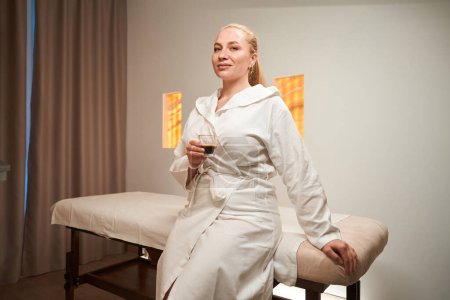 Photo for Blonde female in bathrobe sits in massage room with a drink in her hands, she sat down on massage table - Royalty Free Image