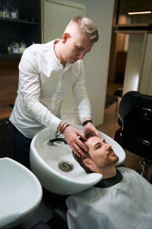 Foto de Joven sonriente disfrutando del proceso de lavarse el cabello en una peluquería, un peluquero trabaja en un lugar de trabajo bien equipado - Imagen libre de derechos