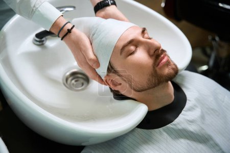 Téléchargez les photos : Coiffeur essuie les cheveux du client après le lavage avec une serviette douce, le maître travaille sur un lieu de travail bien équipé - en image libre de droit