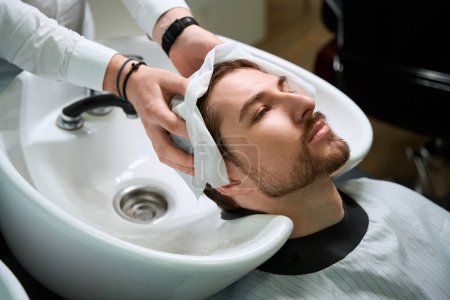 Foto de Maestro limpia el cabello de los clientes después de lavarse con una toalla suave, el barbero trabaja en un lugar de trabajo bien equipado - Imagen libre de derechos
