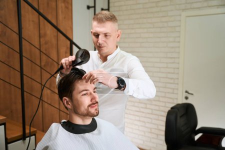 Foto de Master pone el cabello al cliente con un peine y secador de pelo, interior moderno en la barbería - Imagen libre de derechos