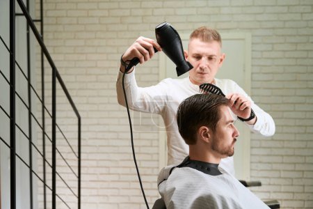 Foto de Peluquero masculino en el trabajo en el lugar de trabajo, hace peinado de pelo a un hombre joven - Imagen libre de derechos