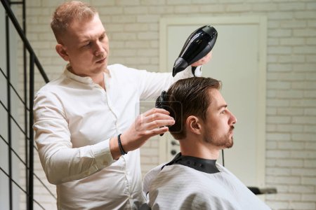 Photo for Hairdresser at work in a well-lit workplace, he is doing hair styling to a young male - Royalty Free Image