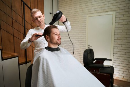 Foto de Peluquería detrás de las obras con un secador de pelo y peine en el lugar de trabajo, que hace el peinado a un joven varón - Imagen libre de derechos