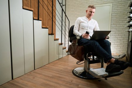 Foto de El hombre con una camisa ligera se sienta en una silla de peluquería, tiene un portátil y un teléfono en sus manos - Imagen libre de derechos