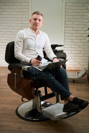 Photo for Man is located in a comfortable hairdressing chair, he uses a laptop and a mobile phone - Royalty Free Image