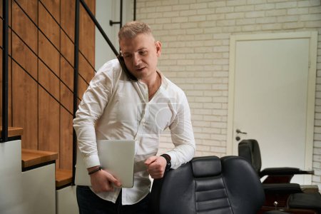 Téléchargez les photos : Jeune homme communique sur un téléphone portable à l'intérieur dans un salon de coiffure, il a un ordinateur portable dans ses mains - en image libre de droit