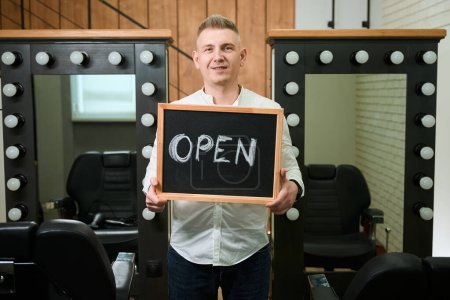 Téléchargez les photos : Homme avec un panneau ouvert se tient dans le salon de coiffure aux miroirs et confortables chaises de coiffure - en image libre de droit