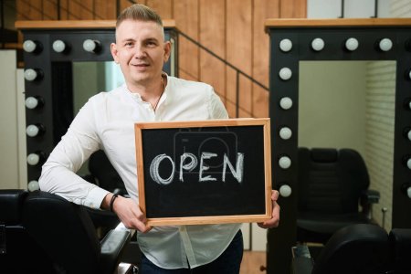 Téléchargez les photos : Homme blond avec un panneau ouvert se tient dans le salon de coiffure aux miroirs et confortables chaises de coiffure - en image libre de droit
