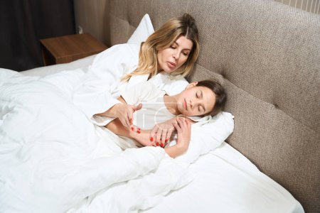 Téléchargez les photos : Femme en manteau blanc et sa fille se relaxant sur le lit dans la chambre d'hôtel - en image libre de droit