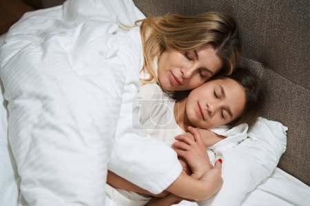 Téléchargez les photos : Mère couchée au lit près de la fille et l'embrassant dans la chambre d'hôtel - en image libre de droit