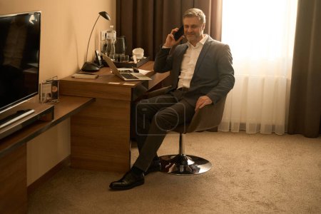 Téléchargez les photos : Homme adulte en costume d'affaires assis sur la chaise au travail et appelant sur smartphone dans le motel - en image libre de droit