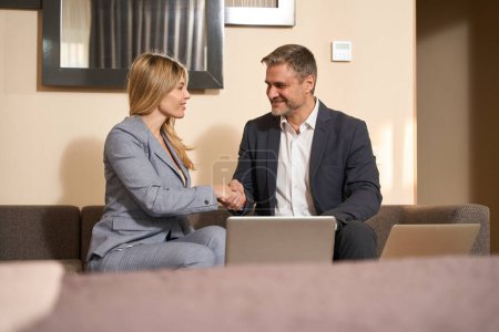 Photo for Man and woman in business suits sitting on sofa and looking at each other, shaking hands in the motel - Royalty Free Image