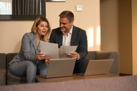 Photo for Adult man and smiling woman in business suits sitting on sofa and working, discussion documents in the motel - Royalty Free Image
