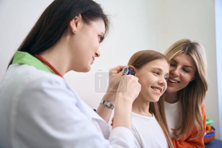 Photo for ENT specialist using otoscope with light to check ear to girl patient, little girl smiling, satisfied with painless medical check-up - Royalty Free Image