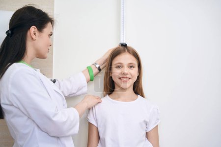 Téléchargez les photos : Infirmière vérifiant la hauteur de la petite fille à l'aide d'une planche à mesurer spéciale sur le mur, bilan de santé des élèves avant l'école - en image libre de droit
