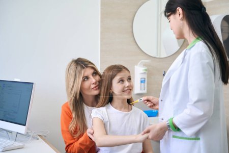 Photo for Friendly woman pediatrist giving thermometer to little girl sitting on mothers knees to check the body temperature, helth check-up stage - Royalty Free Image