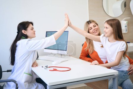 Photo for Qualified woman pediatrist exchanging high-five with little girl patient who sitting on mother knees, satisfied with health check-up in private clinic - Royalty Free Image