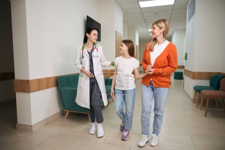 Photo for Cheerful pediatrician showing the clinic building to woman and her daughter patient before complex health check-up, girl will go to rehabilitation - Royalty Free Image