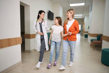 Foto de Terapeuta siguiendo con la mujer y su hija a la sala de examen a lo largo de largo corredor de la clínica privada, médico explicando las etapas de chequeo de la salud - Imagen libre de derechos
