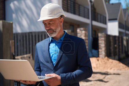 Téléchargez les photos : Géodésie masculine en costume d'affaires et casque de sécurité inspectant le chantier, arpentant les terres tenant l'ordinateur portable dans les mains, examinant le paysage - en image libre de droit