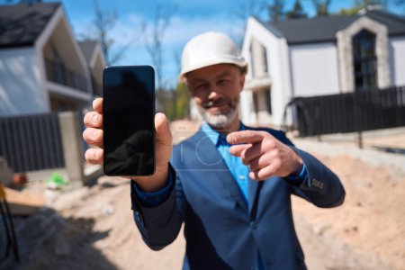 Téléchargez les photos : Investisseur barbu d'immeuble d'habitation tenant smartphone, montrant une plate-forme de négociation électronique en ligne, appel d'offres de vente de maison - en image libre de droit