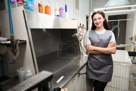 Téléchargez les photos : Femme gaie opérateur de nettoyage à sec debout près de la table à repasser et souriant, service de blanchisserie industrielle - en image libre de droit