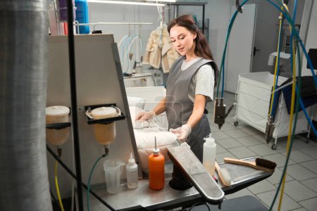 Foto de Operadora femenina de servicio de limpieza en seco inspeccionando el artículo, haciendo tratamiento previo antes de la limpieza, industria de lavandería profesional - Imagen libre de derechos