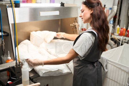 Téléchargez les photos : Opérateur de nettoyage à sec femme vue de côté inspectant l'article après la procédure de nettoyage, post-traitement - en image libre de droit