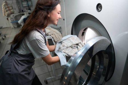 Téléchargez les photos : Femme service de blanchisserie travailleur chargement grande machine à laver professionnelle avec des vêtements, tissus délicats nettoyage à sec - en image libre de droit