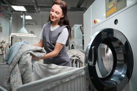 Foto de Mujer feliz servicio de limpieza en seco trabajador carga lavadora industrial con ropa, limpieza con solución sin agua, lavandería profesional - Imagen libre de derechos