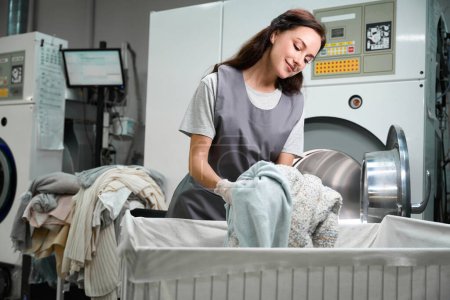 Téléchargez les photos : Lady travailleur de service sec enlever les vêtements lavés de la grande machine et de mettre à la poubelle, post-traitement - en image libre de droit