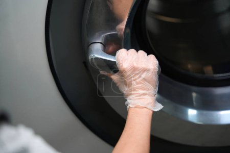 Téléchargez les photos : Femme en gants de protection tenant une grande poignée de machine à laver industrielle, ouvrant la porte pour charger ou enlever les vêtements, service de blanchisserie - en image libre de droit