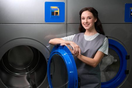 Foto de Mujer atractiva trabajadora de servicios de limpieza en seco de pie cerca de una gran máquina industrial para lavar o secar, instalaciones profesionales - Imagen libre de derechos