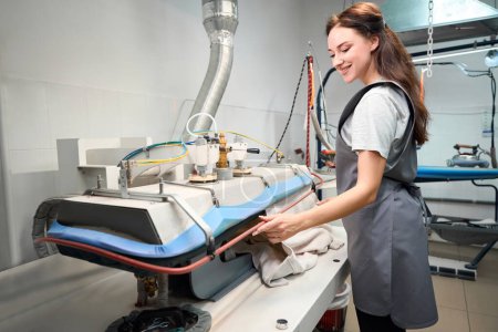 Téléchargez les photos : Femme opérateur de bureau de nettoyage à sec repassage des vêtements avec presse à vapeur spéciale pour lisser et façonner les vêtements - en image libre de droit