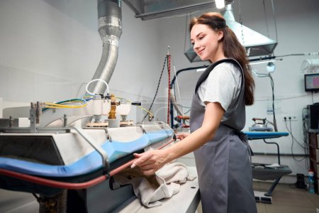 Foto de Mujer sonriente de limpieza en seco trabajador de oficina secado y planchado de ropa con prensa de vapor, instalaciones profesionales - Imagen libre de derechos