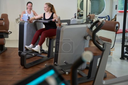 Photo for Joyful client of clinic is engaged in a modern fitness machine, a trainer consultant is nearby - Royalty Free Image